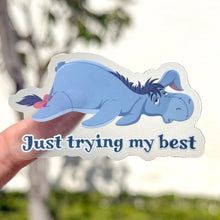 Load image into Gallery viewer, Sticker of a blue eeyore with text 'Just trying my best' held by a hand against a blurred natural background.
