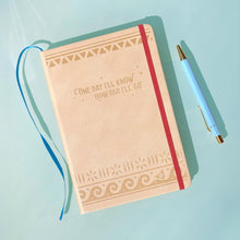 Load image into Gallery viewer, A beige hardcover notebook with gold tribal-style border designs and the quote 'COME DAY I'LL KNOW HOW YOU'LL GO' on the front, shown with a blue ribbon bookmark and a light blue pen on a light blue background.
